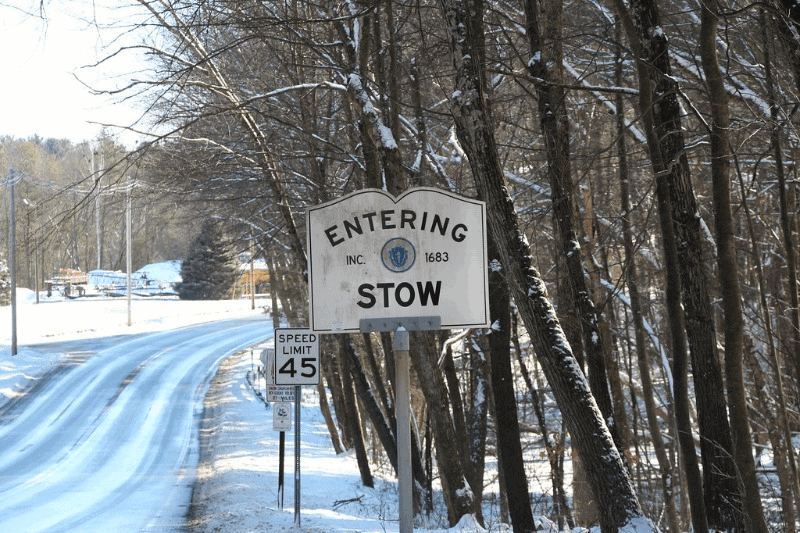 Road sign indicating the entrance to Stow, a town established in 1683, surrounded by a snowy landscape with trees lining the road.